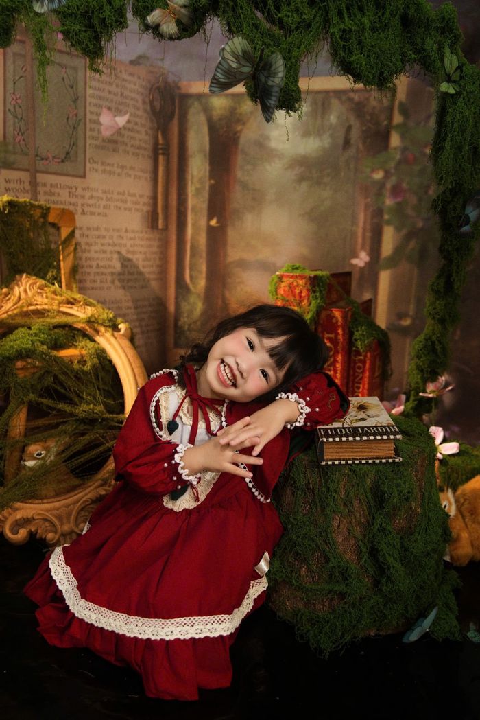 Young girl in a fairytale setting wearing a red dress, surrounded by books and greenery.